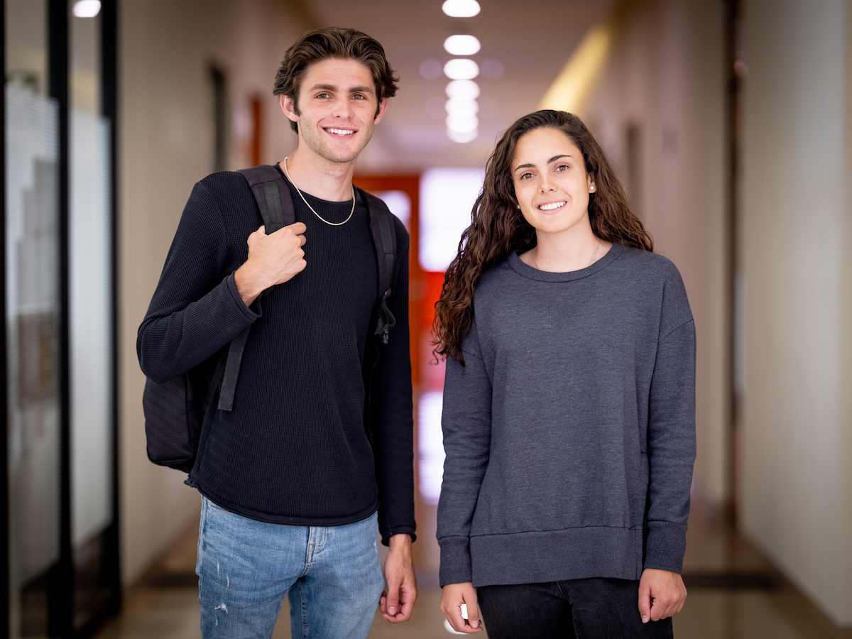 Dos estudiantes sonrientes de pie en un pasillo interior iluminado; él con mochila al hombro y ambos con ropa casual oscura.